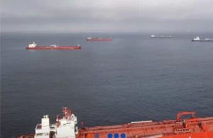 تطفو العديد من سفن الشحن الكبيرة على مياه هادئة تحت سماء ملبدة بالغيوم، ويظهر سطح سفينة حمراء وبيضاء في المقدمة.