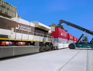 قطار شحن يحمل علامة الاتحاد للقطارات محمل بحاويات شحن حمراء بينما تقوم رافعة شوكية بلون أزرق مخضر بتحميل أو تفريغ الحاويات في يوم صافٍ ومشمس.