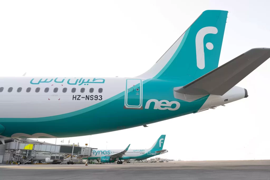 طائرة فلاي ناس بيضاء ومزرقّة اللونين متوقفة عند بوابة المطار، وتظهر زعنفة الذيل وجزء من جسم الطائرة. تظهر طائرات أخرى تابعة لطيران ناس في الخلفية على مدرج المطار.