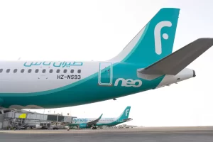 طائرة فلاي ناس بيضاء ومزرقّة اللونين متوقفة عند بوابة المطار، وتظهر زعنفة الذيل وجزء من جسم الطائرة. تظهر طائرات أخرى تابعة لطيران ناس في الخلفية على مدرج المطار.