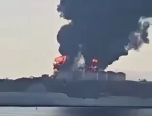 تتصاعد أعمدة كبيرة من الدخان الأسود وألسنة اللهب من منشأة صناعية بالقرب من مسطح مائي، مما يشير إلى نشوب حريق أو انفجار كبير. السماء ضبابية بسبب الدخان.