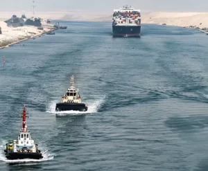 زورقا قطر يسيران على طول قناة واسعة باتجاه سفينة شحن كبيرة محملة بحاويات شحن ملونة. تصطف الضفاف الرملية على جانبي الممر المائي تحت سماء ضبابية.