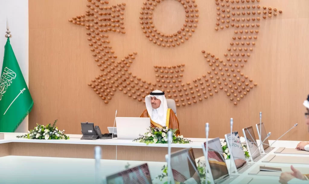 رجل يرتدي الزي السعودي التقليدي يجلس على طاولة مؤتمرات مع أجهزة كمبيوتر محمولة وميكروفونات، أمام ديكور حائط خشبي كبير وعلم المملكة العربية السعودية. يبدو المكان رسمياً ورسمياً.