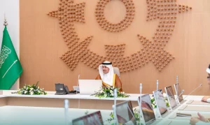 رجل يرتدي الزي السعودي التقليدي يجلس على طاولة مؤتمرات مع أجهزة كمبيوتر محمولة وميكروفونات، أمام ديكور حائط خشبي كبير وعلم المملكة العربية السعودية. يبدو المكان رسمياً ورسمياً.