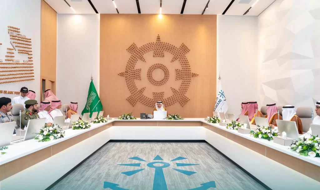 اجتماع رسمي مع المسؤولين الجالسين حول طاولة على شكل حرف U، تظهر أعلام المملكة العربية السعودية وشعار ترس كبير على الحائط. يحضر الاجتماع رجال ونساء على حد سواء، ويرتدي معظمهم ملابس شرق أوسطية تقليدية.