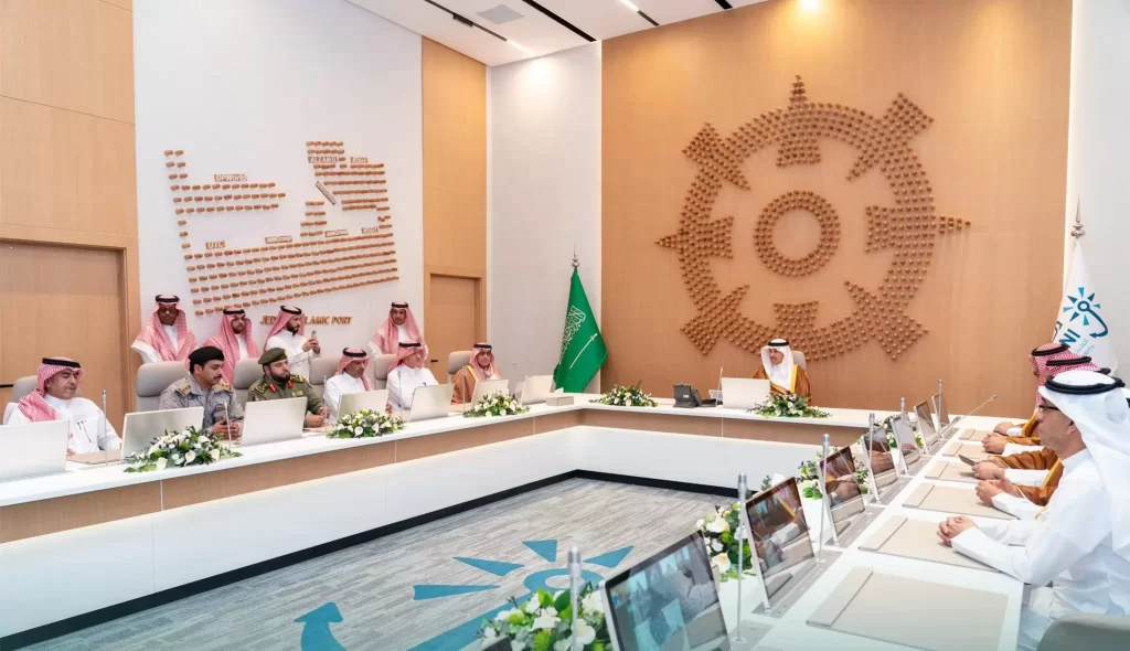 تجلس مجموعة من الرجال بالزي السعودي التقليدي وبدلات رجال الأعمال حول طاولة اجتماعات حديثة مع أجهزة كمبيوتر محمولة؛ وتظهر الأعلام السعودية وشعار حائط كبير في الغرفة.